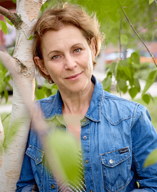 Helen in a blue denim shirt stands in the greenery outdoors. Half-length photo.
