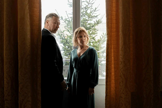 Helen Sjöholm i grön klänning och Mikael Persbrandt i mörk kostym stående framför ett fönster med långa brungula gardiner.