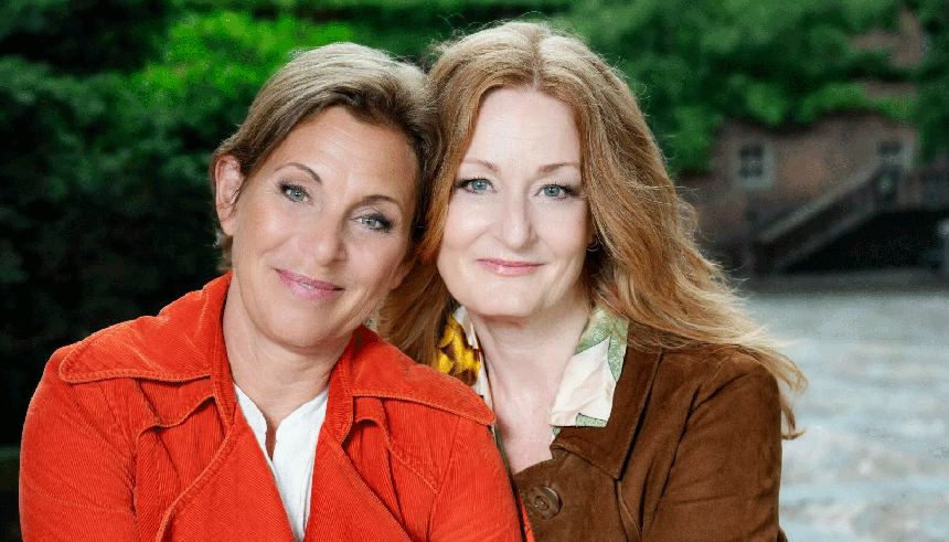Helen Sjöholm i ljusröd kappa och Anna Stadling i brun, sittande med huvudena tillsammans utomhus. Foto i halvfigur.
