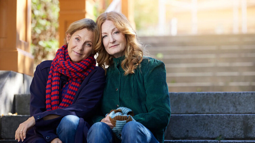 Helen och Anna höstklädda, sittande i en trappa utomhus med huvudena lutade mot varandra.
