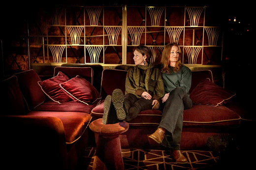 Helen och Anna, klädda i grönt, sitter i röd sammetssoffa. Helen har fötterna på en pall och visar skosulorna.